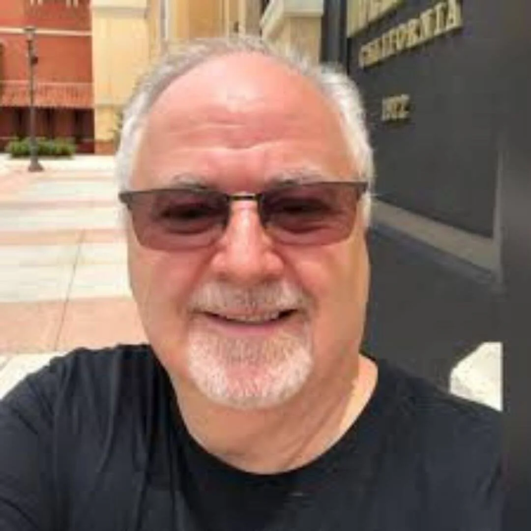 A man with sunglasses smiling outdoors near a building.