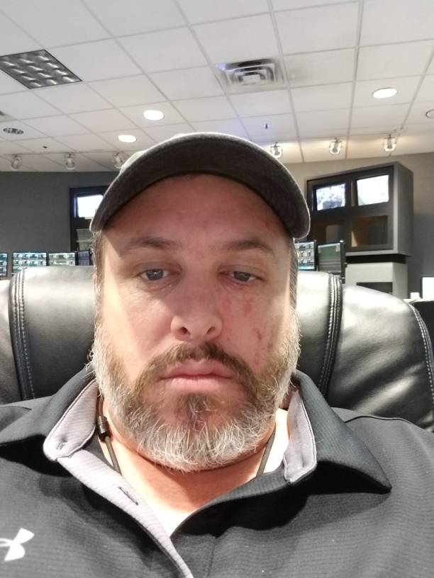 A man with a beard and baseball cap takes a selfie while sitting in a black leather chair.