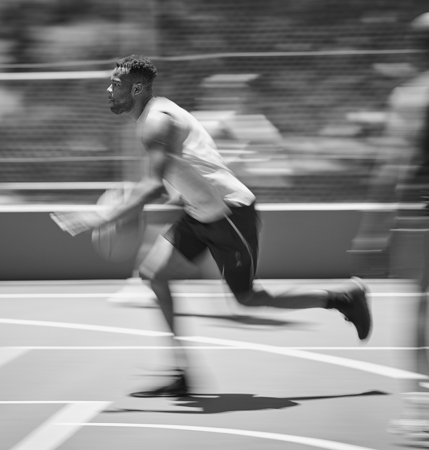 A black and white motion-blur photo of a man running and dribbling a basketball on a court.