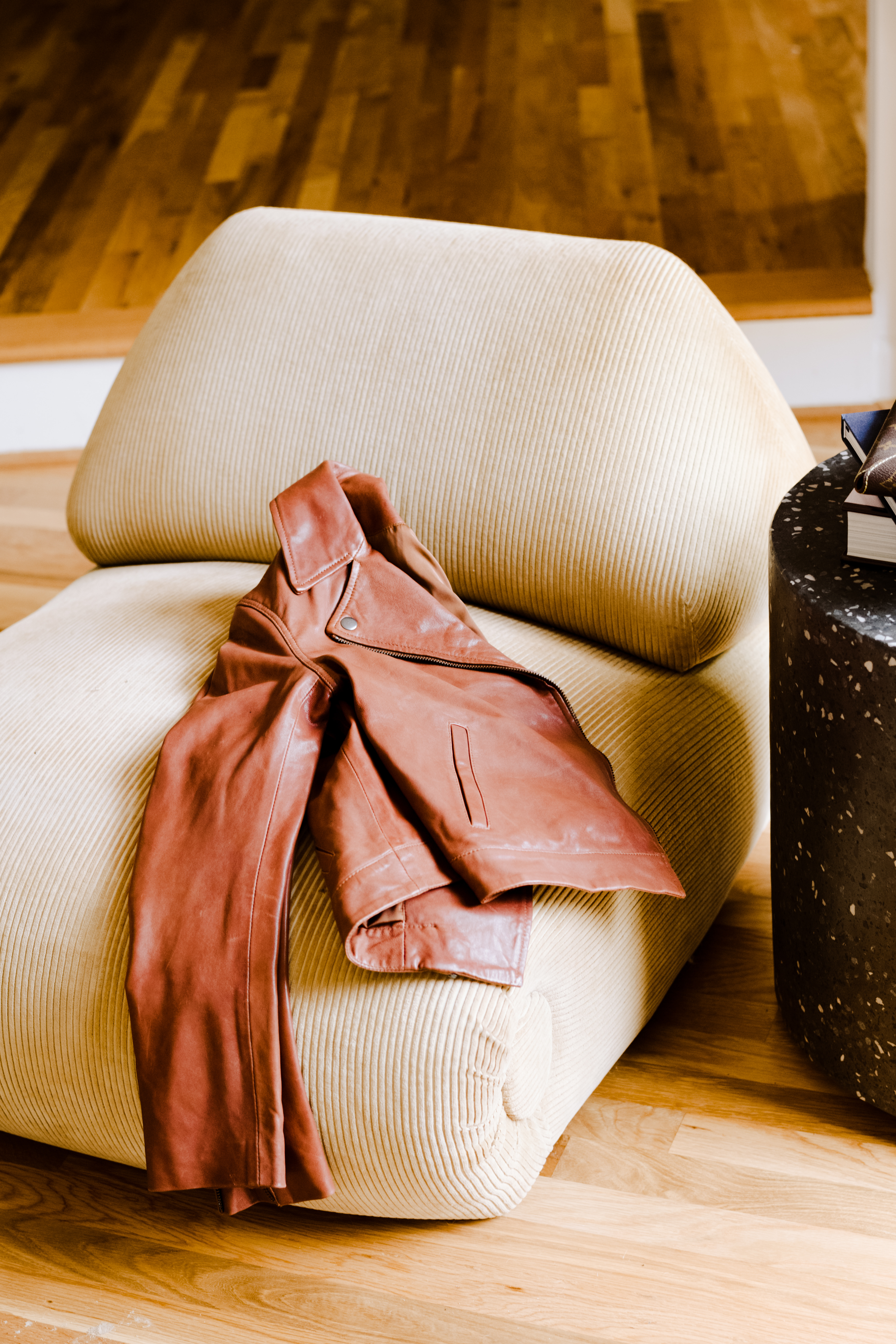 Brown leather jacket draped over a beige chair on wooden floor.