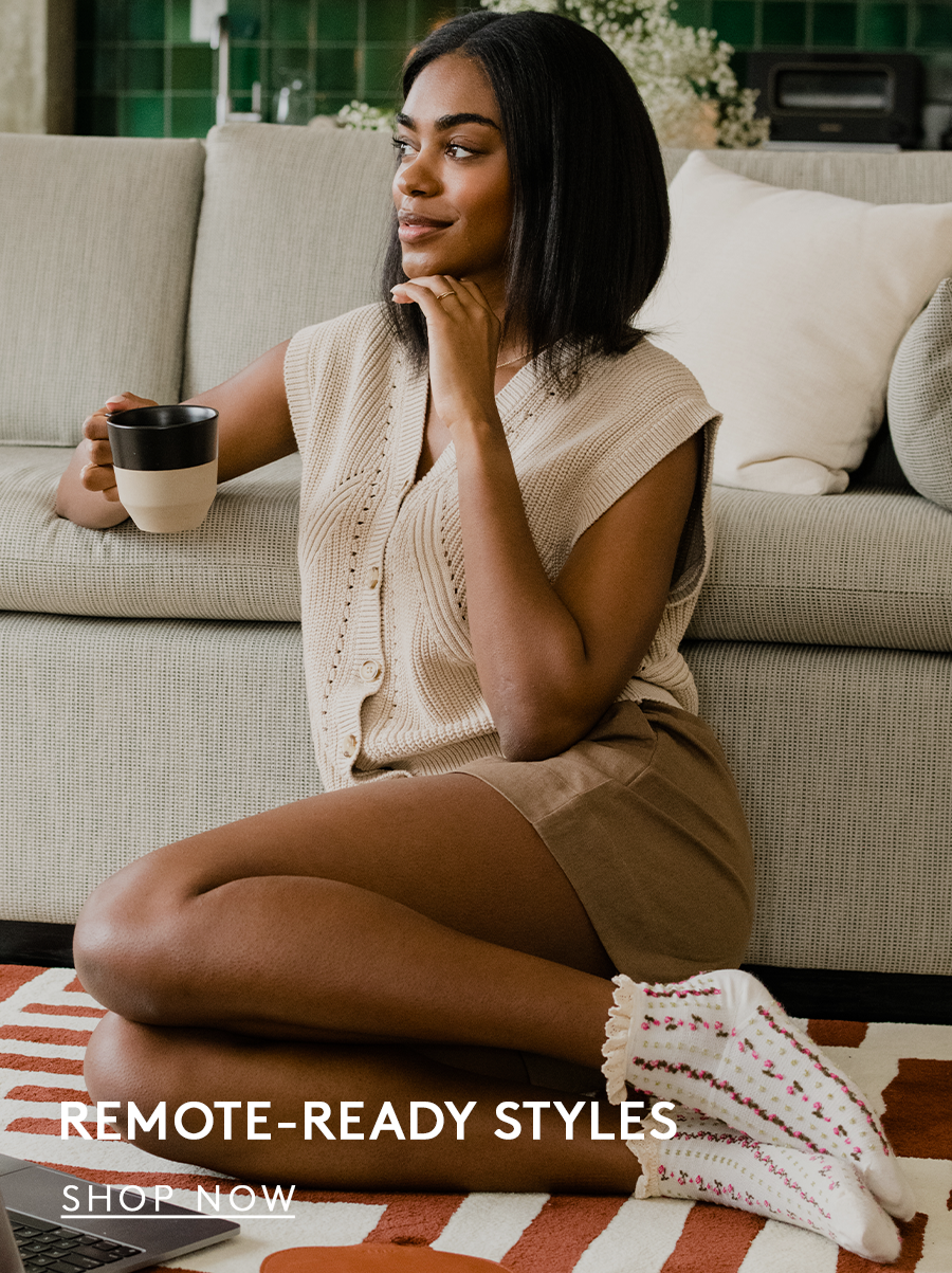 woman sitting on the floor beside a couch with a mug in her hand, wearing a sweater, shorts, and socks text reads remote ready styles, shop now