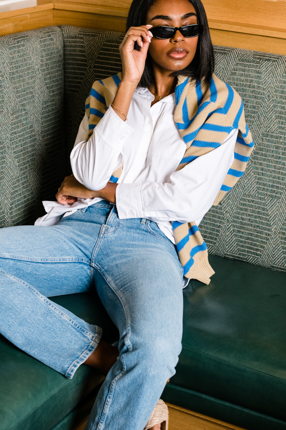 Person sitting stylishly, wearing sunglasses and casual attire with a sweater draped over shoulders.