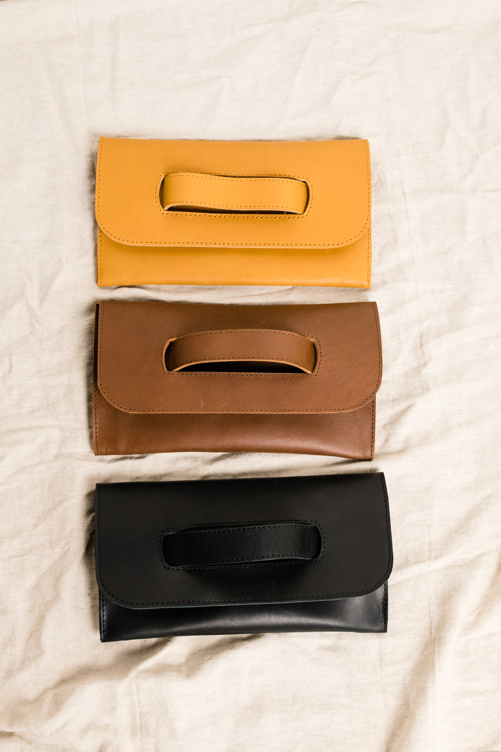 Three leather clutches in yellow, brown, and black on a beige fabric background.