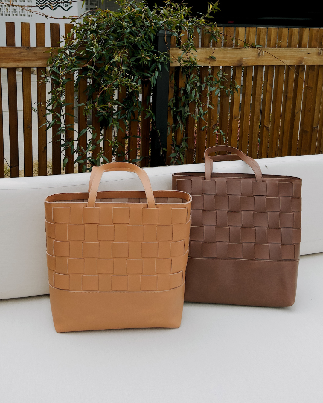 Two woven leather bags on a white surface, with a wooden fence and plants in the background.