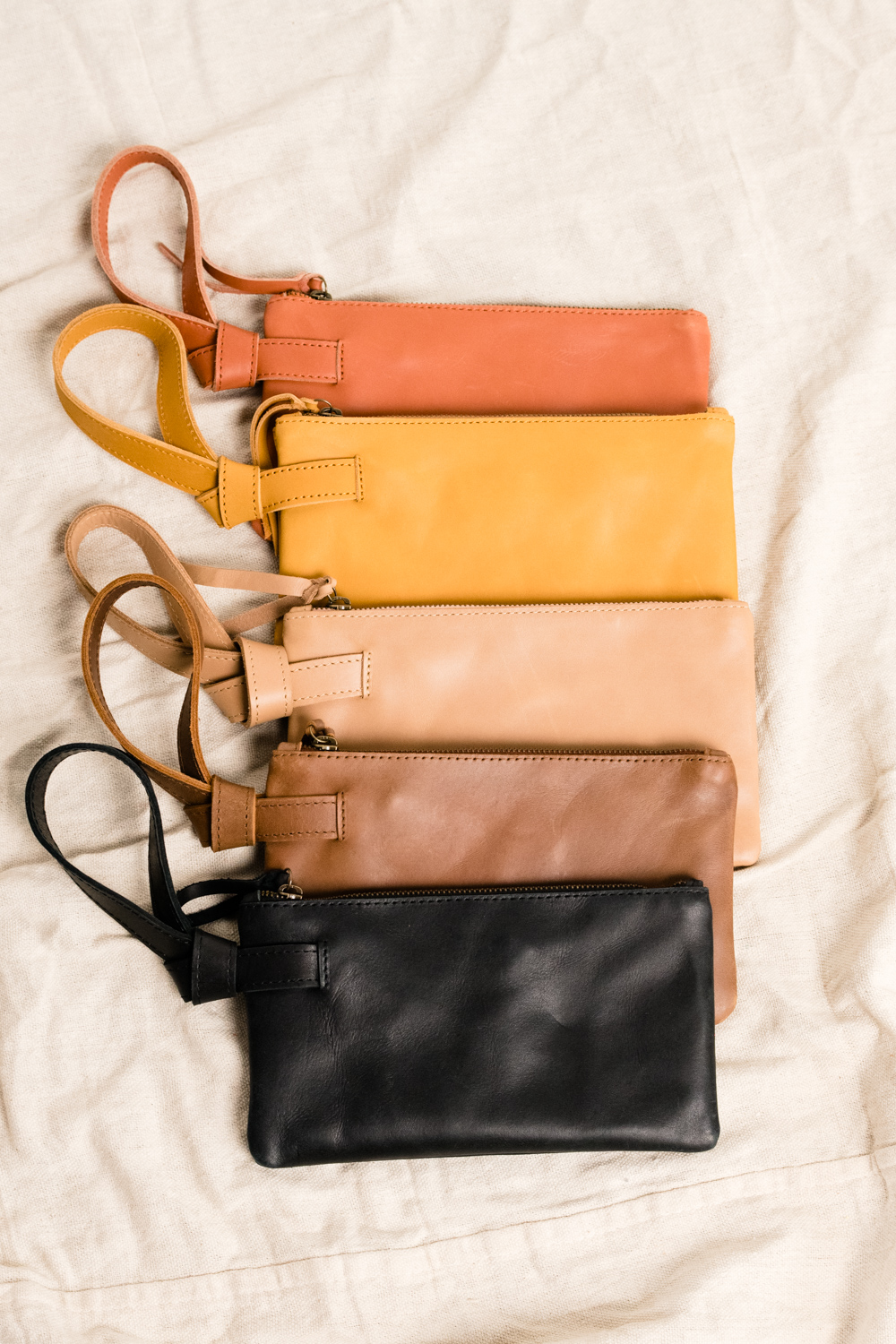 Five leather wristlet pouches in various colors arranged on a beige fabric background.