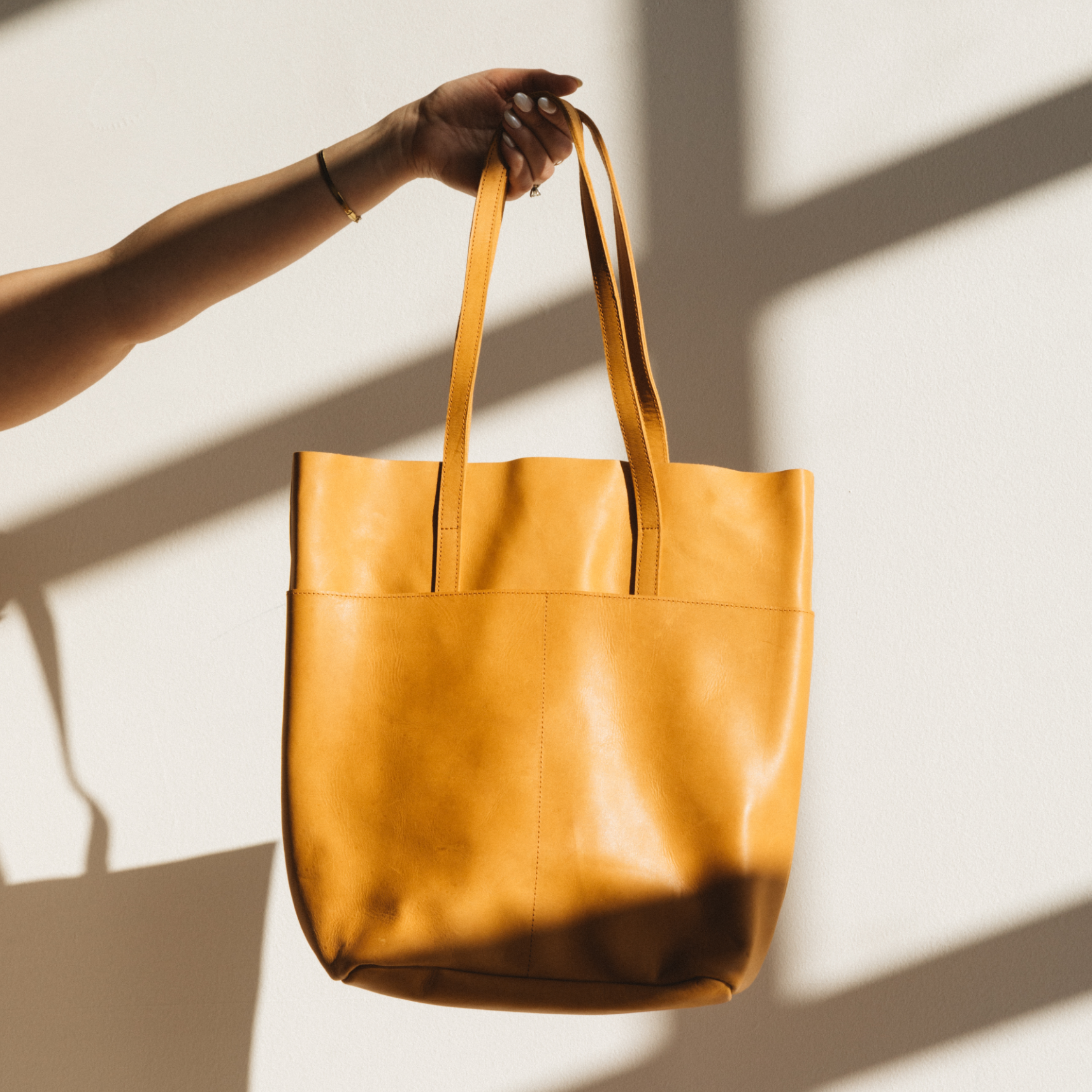 A hand holding a yellow leather tote bag against a wall with shadows.