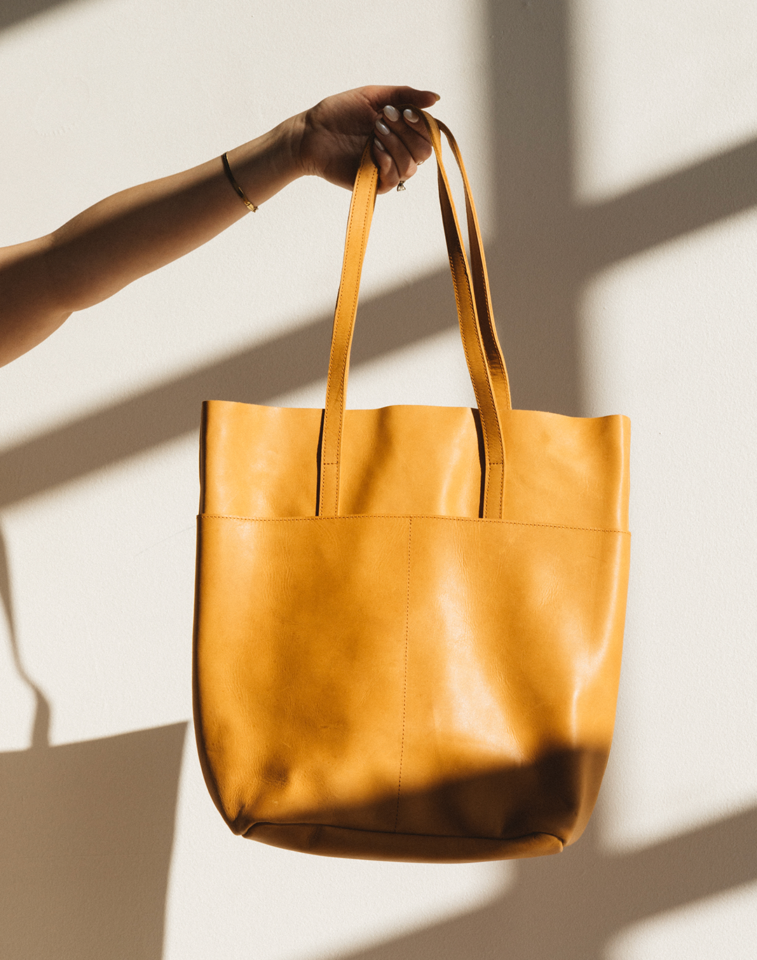 A hand holding a tan leather tote bag against a white wall.