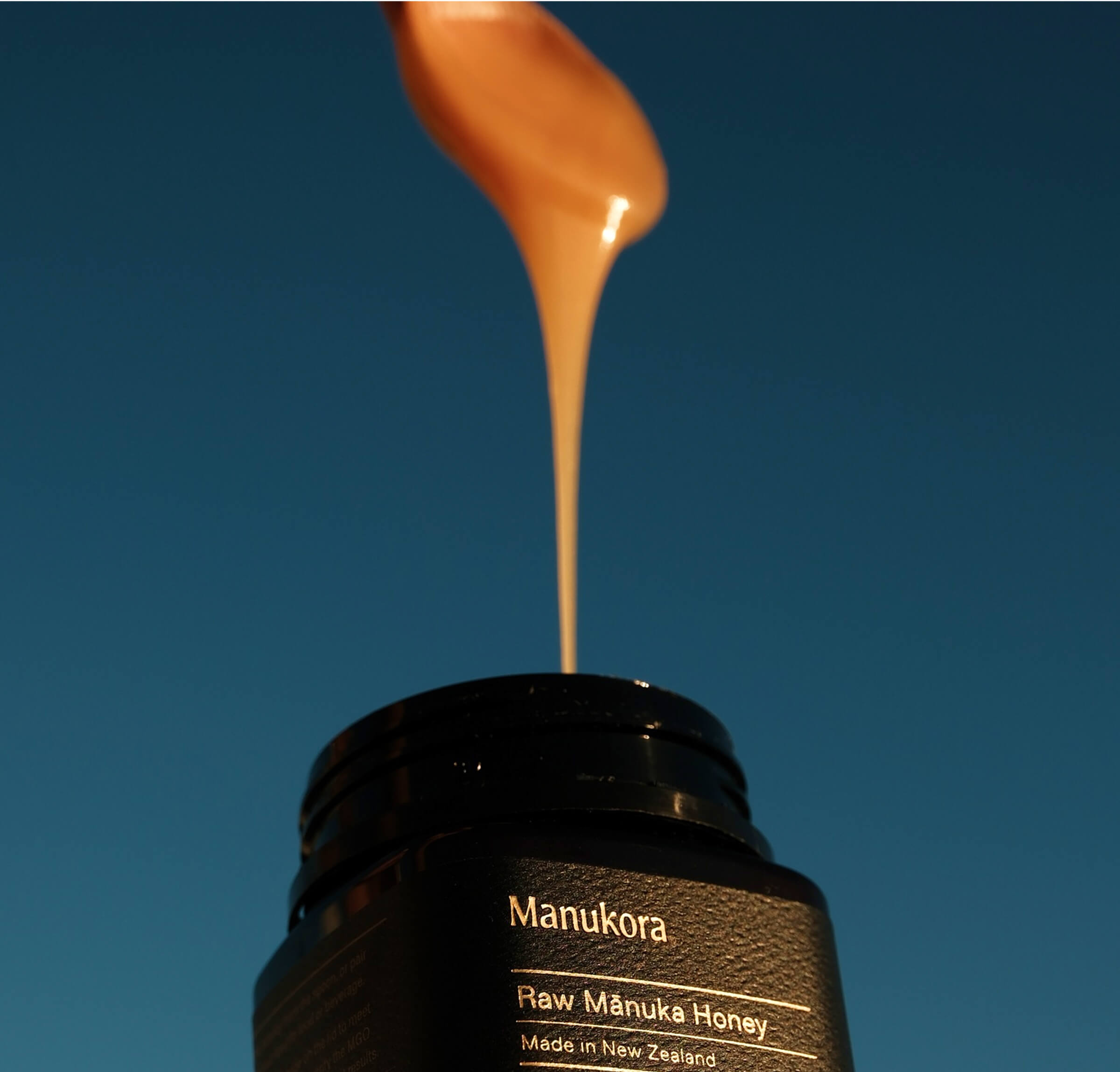 Honey pouring from a spoon into a jar labeled 'Manukora Raw Mānuka Honey.'