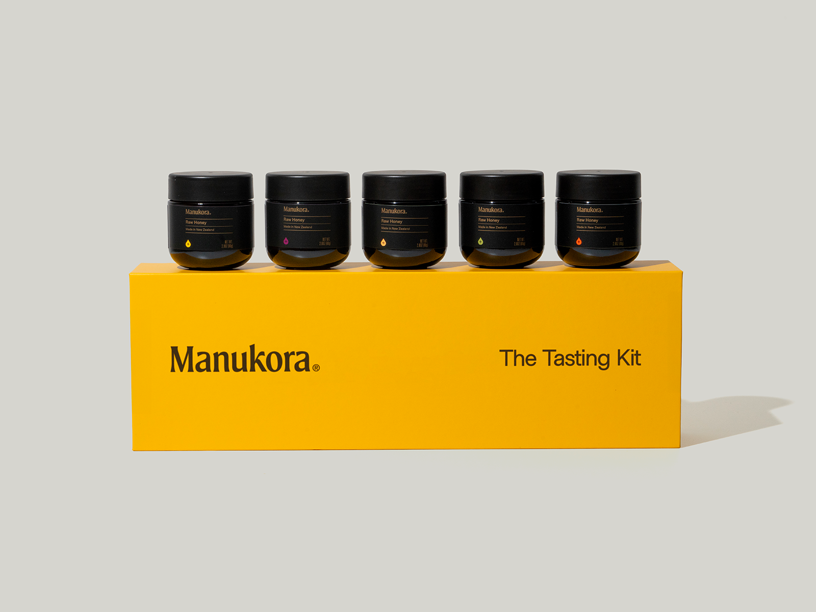 A yellow box labeled 'Manukora The Tasting Kit' with five small black jars of raw honey on top.