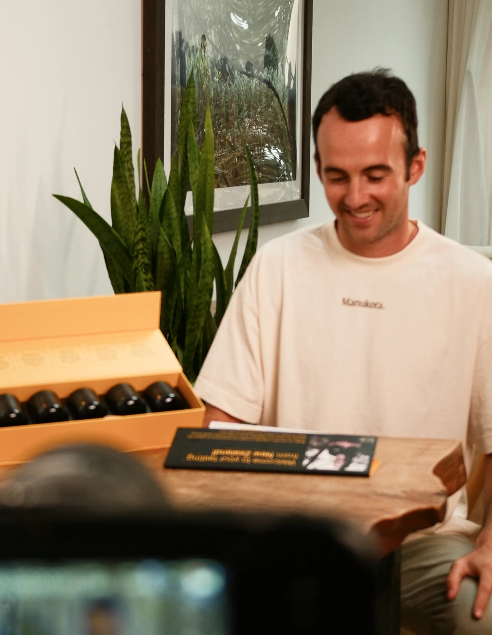 Man sitting at a table with a box of bottles, plants in background.
