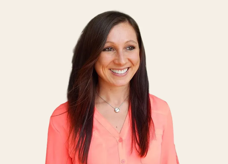 A headshot of a smiling woman with long dark hair, wearing a coral-colored blouse and a necklace.