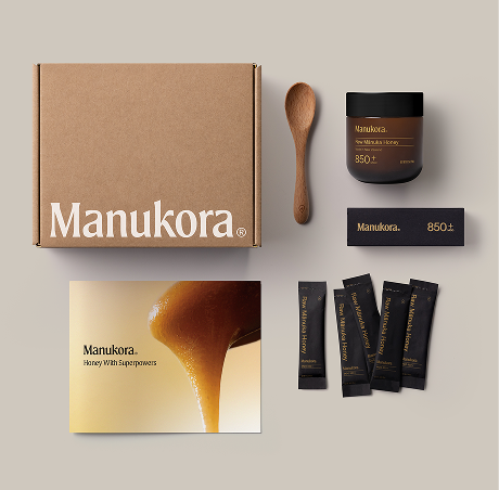 A flat lay of Manukora honey products including a box, a jar, single-serving packets, and a wooden spoon.