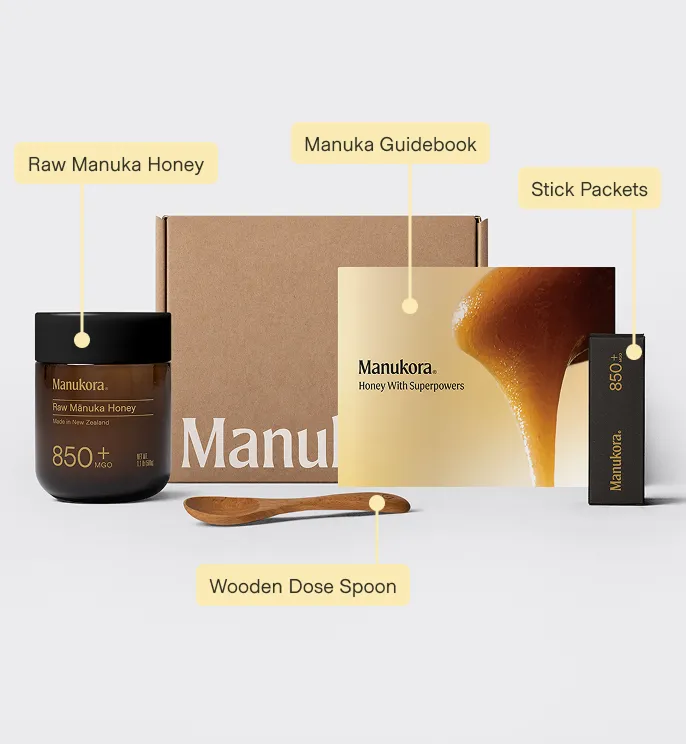 A Manukora honey set with labeled jar, guidebook, stick packets, wooden spoon, and a cardboard box.