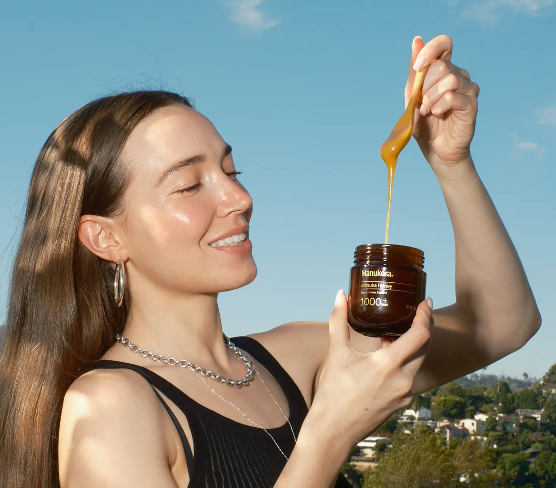 A smiling woman holds a jar of Mānuka honey, with honey dripping from a wooden spoon into it.