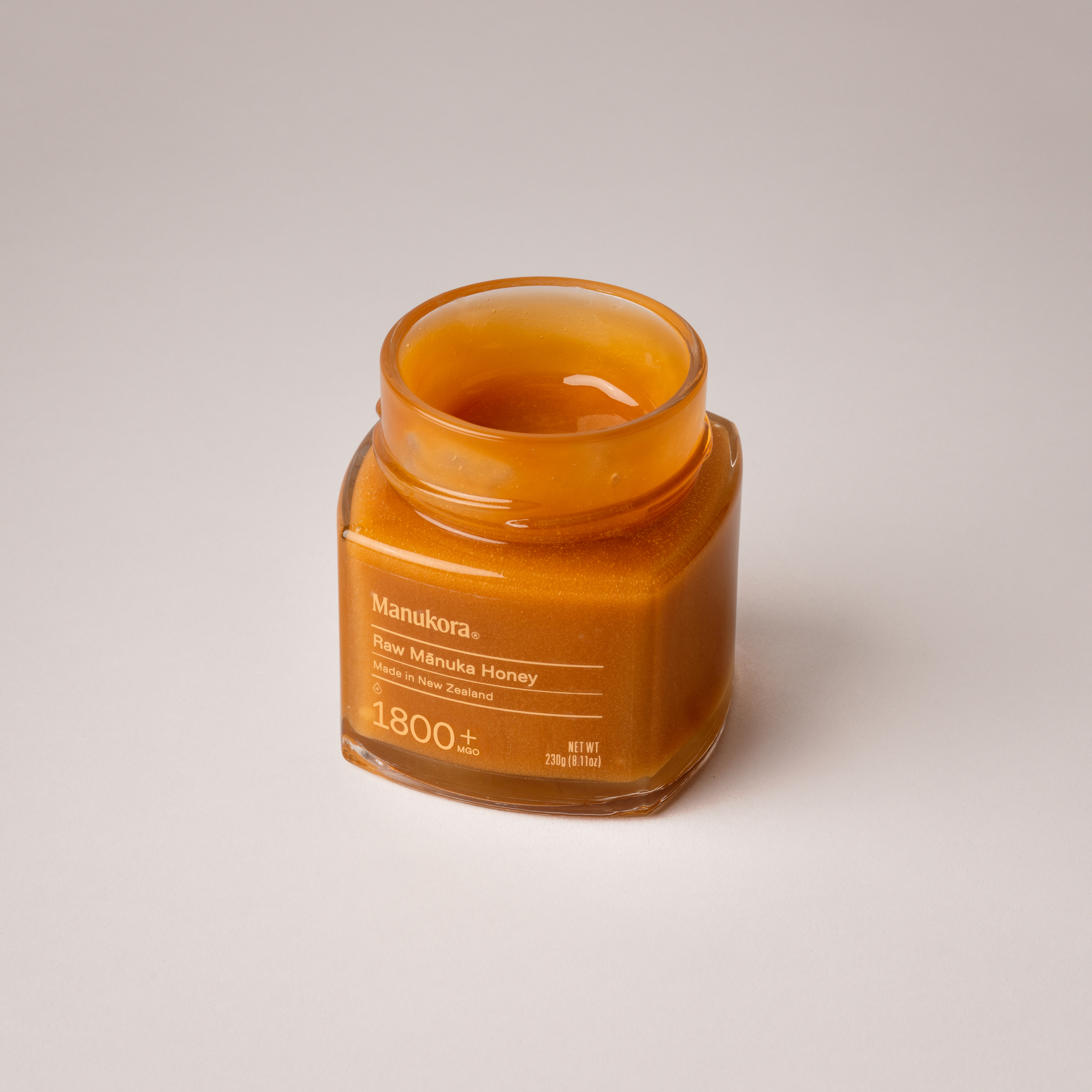 An open jar of Manukora Raw Mānuka Honey against a plain, off-white background.