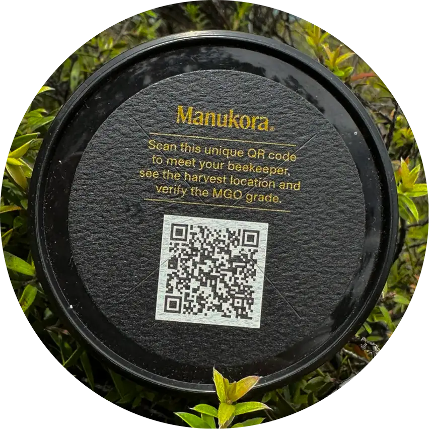 A close-up of a black Manukora jar lid with a QR code, resting on green leaves.