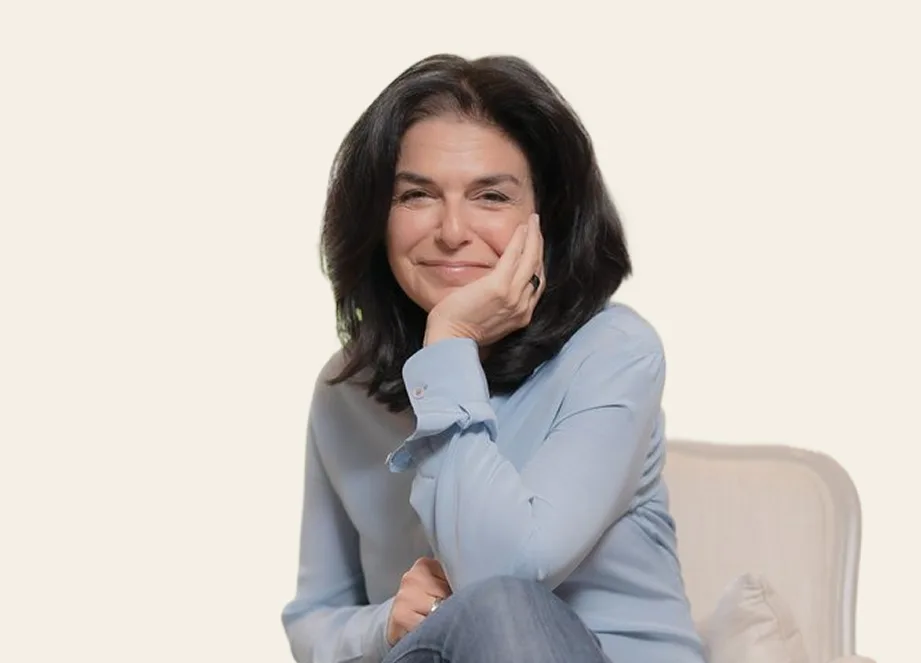 A woman with dark, shoulder-length hair in a light blue shirt smiles, resting her chin on her hand.