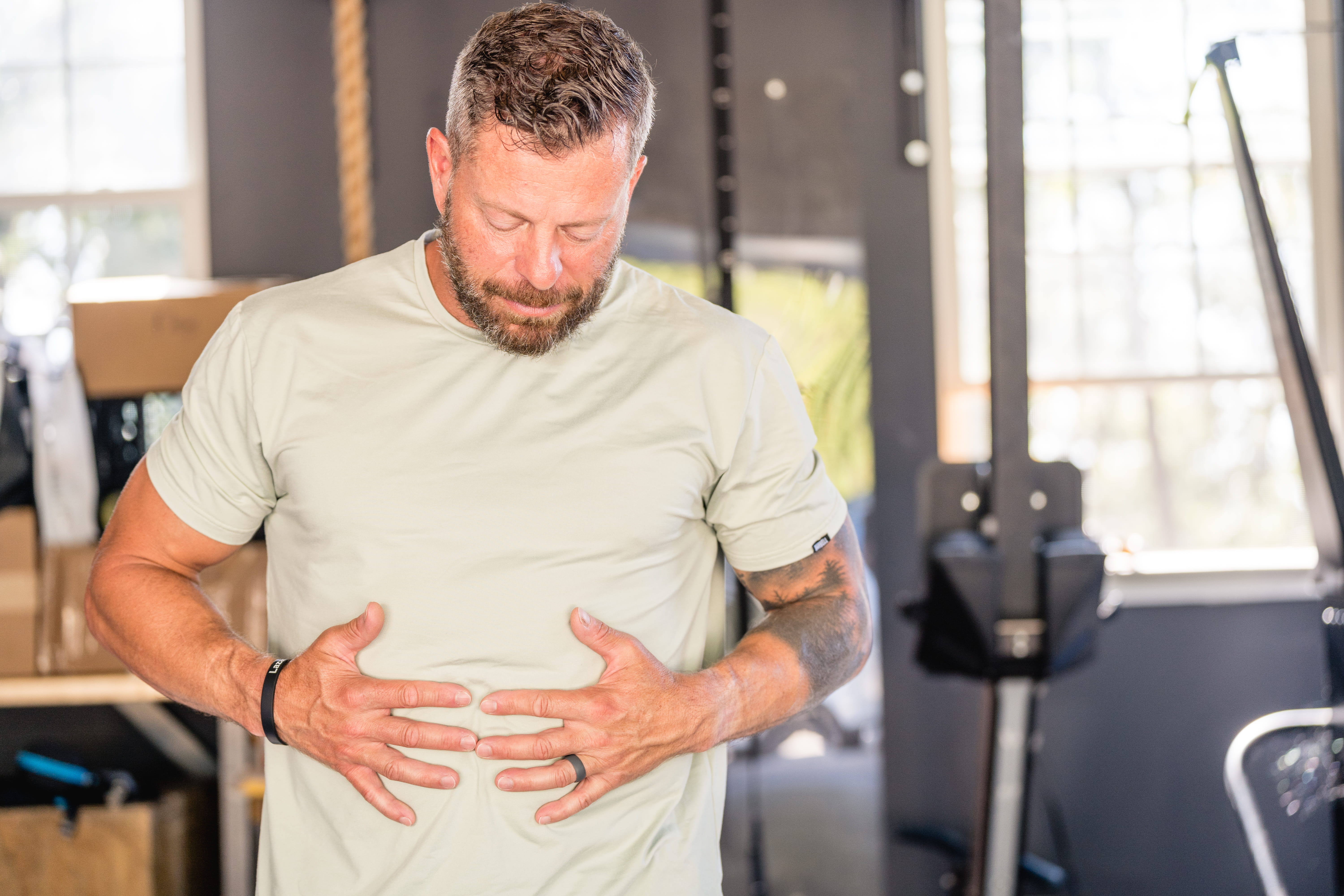 A man in a gym looks down and holds his stomach with both hands, appearing uncomfortable.