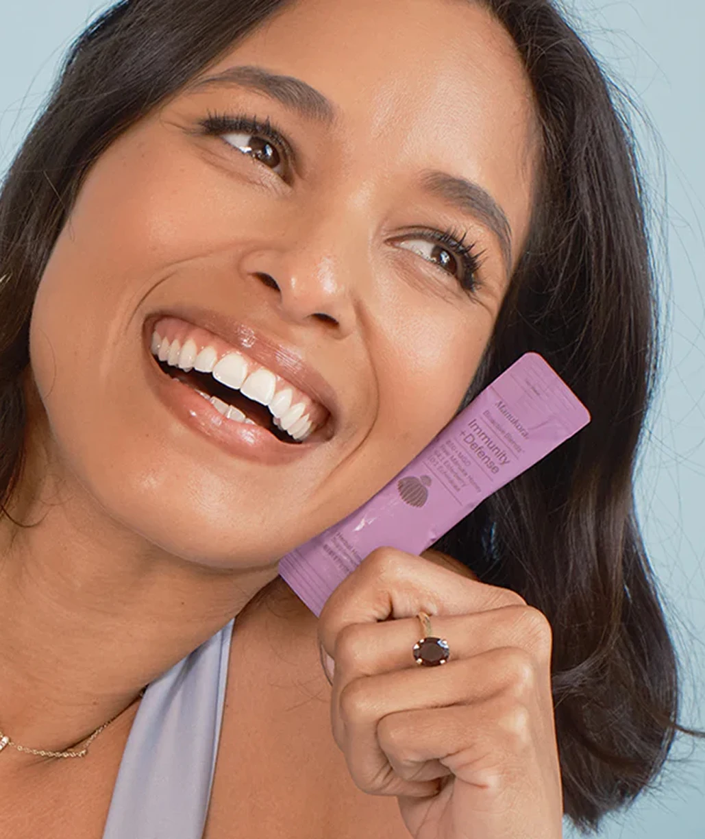 A smiling woman with dark hair holds a purple packet labeled 'Immunity + Defense' against a light blue background.