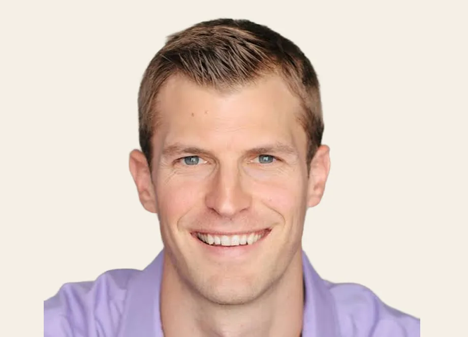 A headshot of a smiling man with short brown hair and blue eyes, wearing a purple shirt.