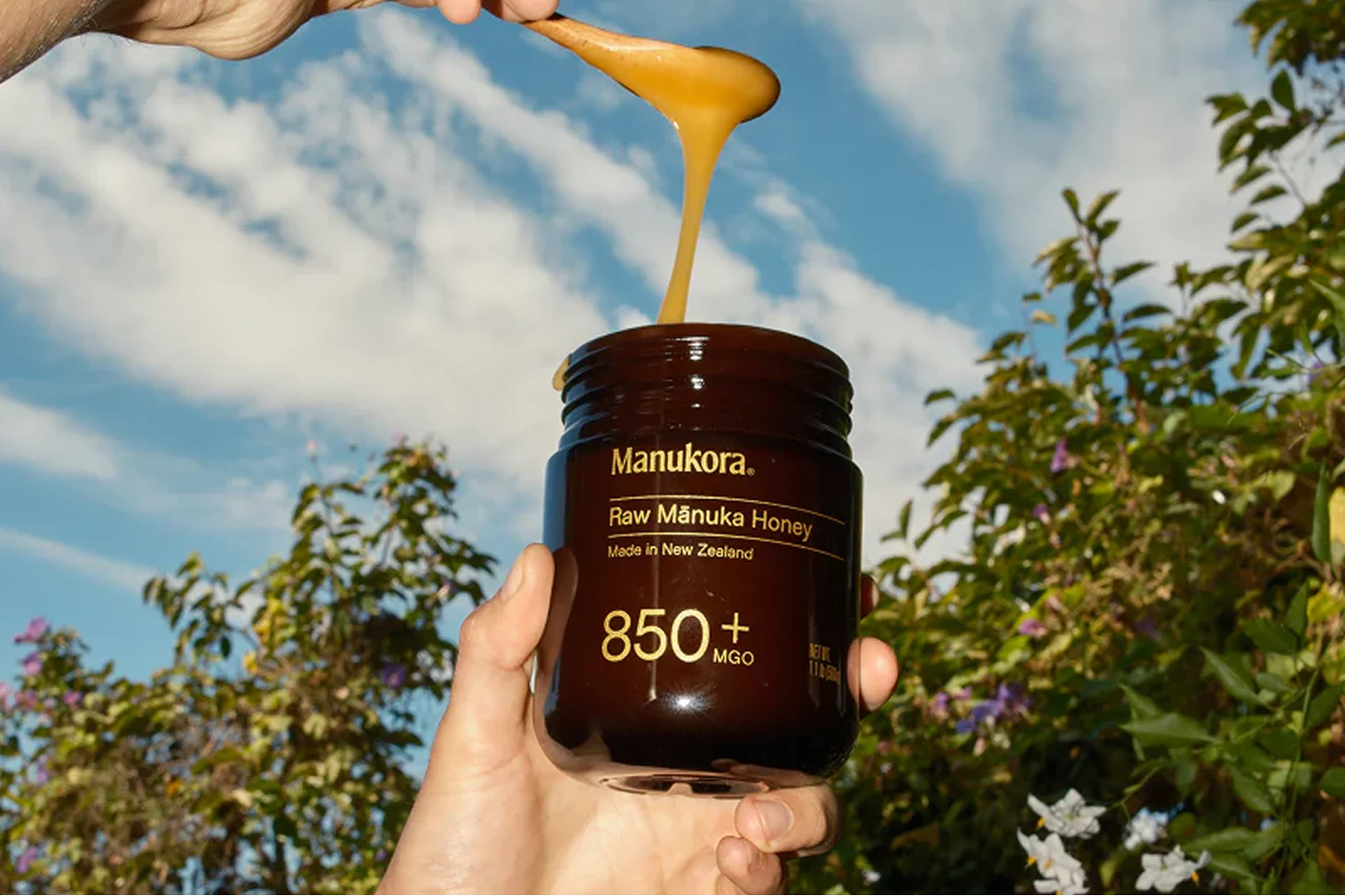 A hand holds a jar of Manukora Raw Mānuka Honey as a spoon drips honey into it.