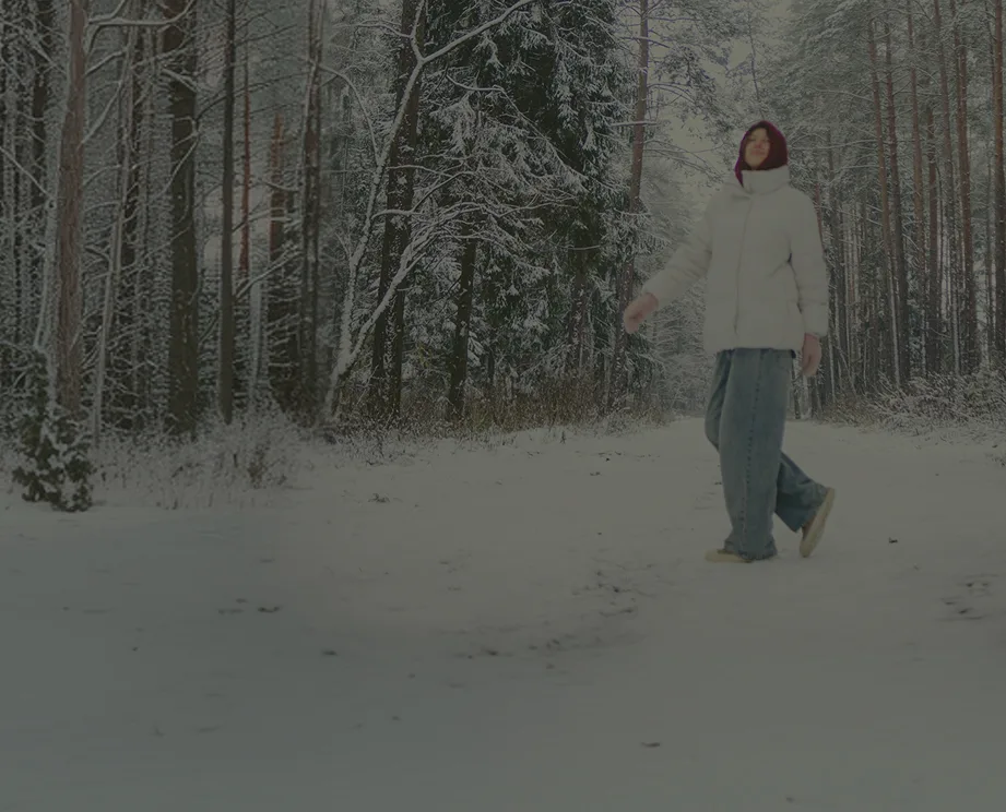 A person wearing a white puffer jacket and red hood walks on a snowy path through a forest.