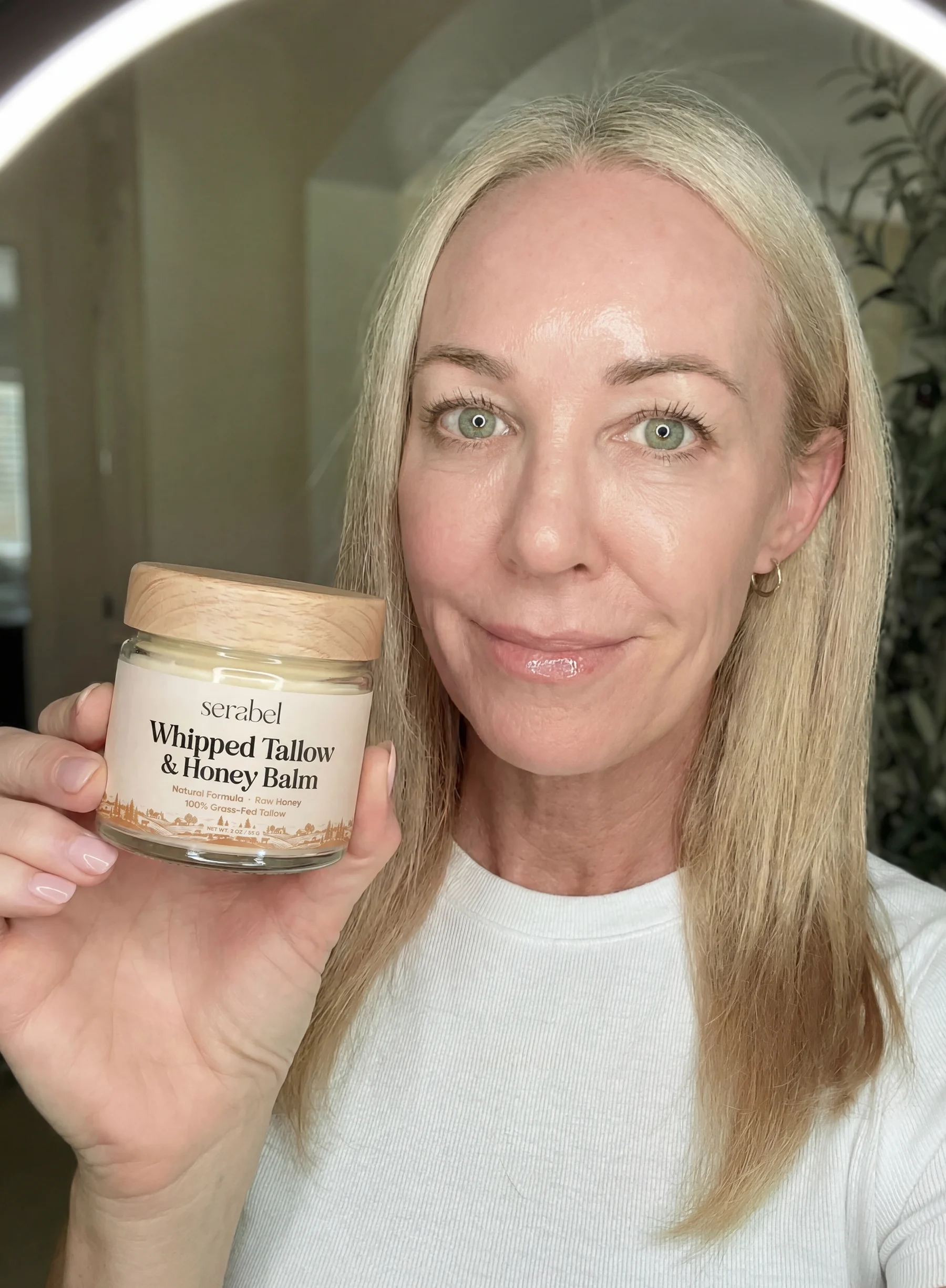 A blonde woman with green eyes holds up a jar of Serabel Whipped Tallow & Honey Balm.