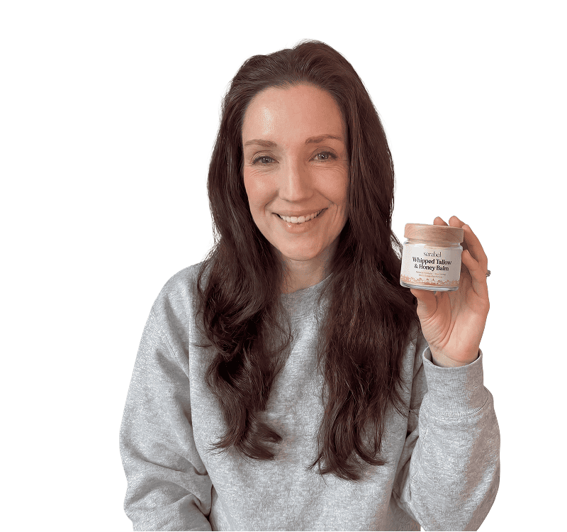 A smiling woman in a grey sweatshirt holds a small jar of Whipped Tallow & Honey Balm.