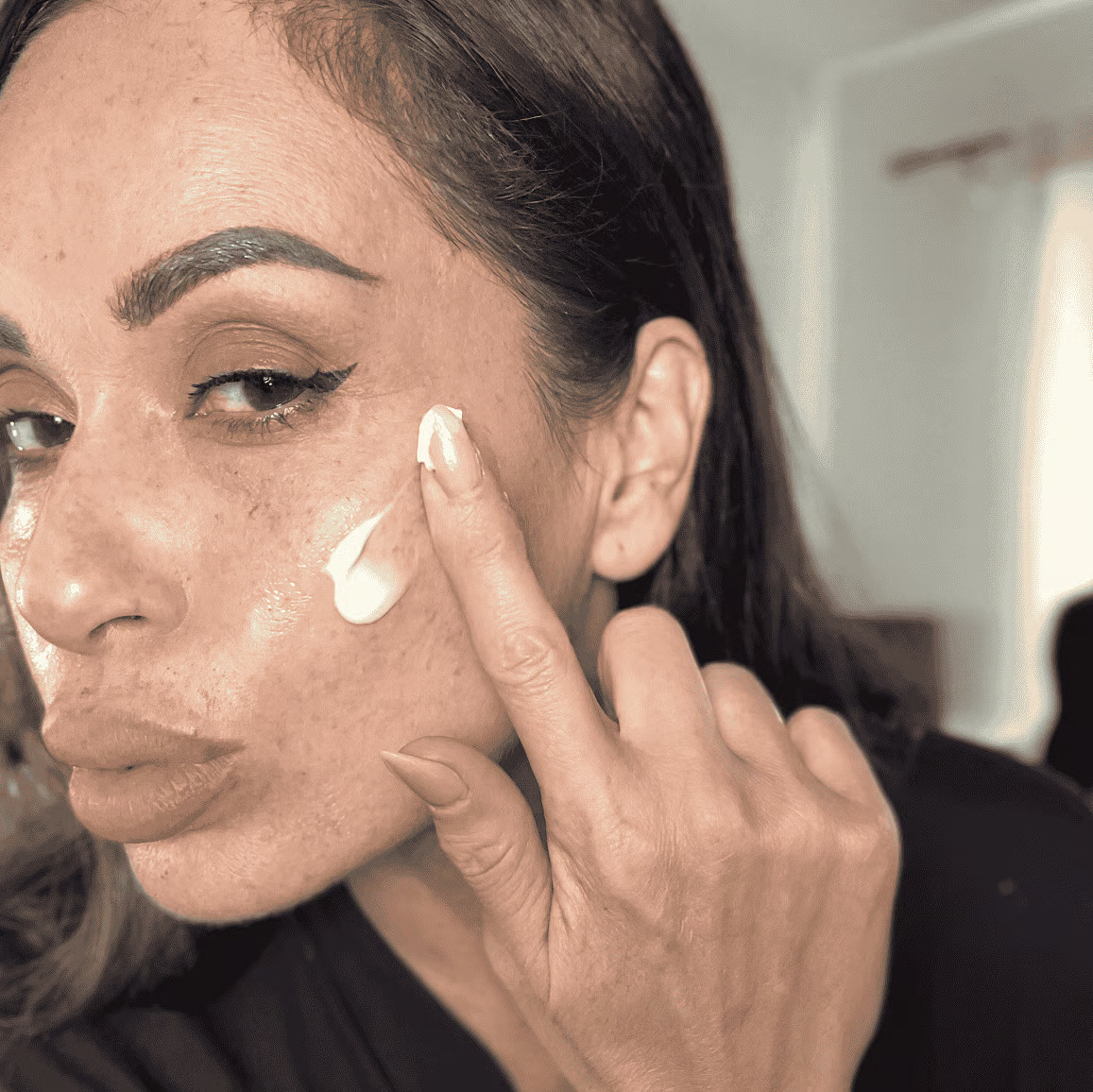 A close-up shot of a woman with eyeliner applying a white cream to her cheek with her finger.