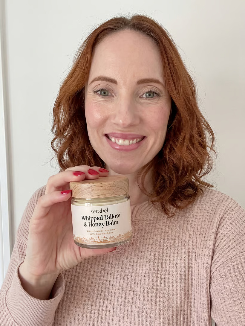 A smiling woman with red hair holds a jar of Serabel Whipped Tallow & Honey Balm.