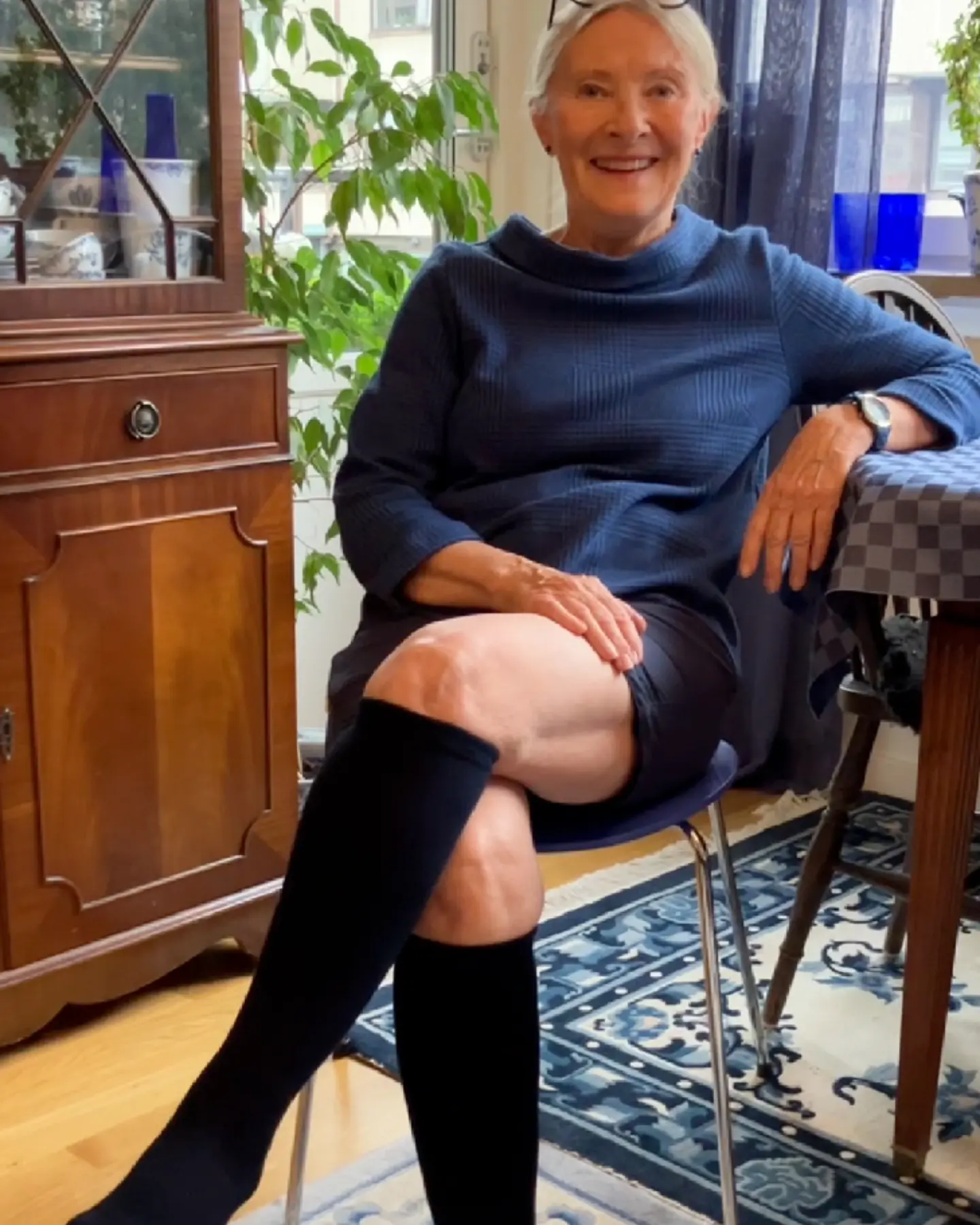 Smiling person sitting indoors with crossed legs, wearing a blue dress and black knee-high socks.