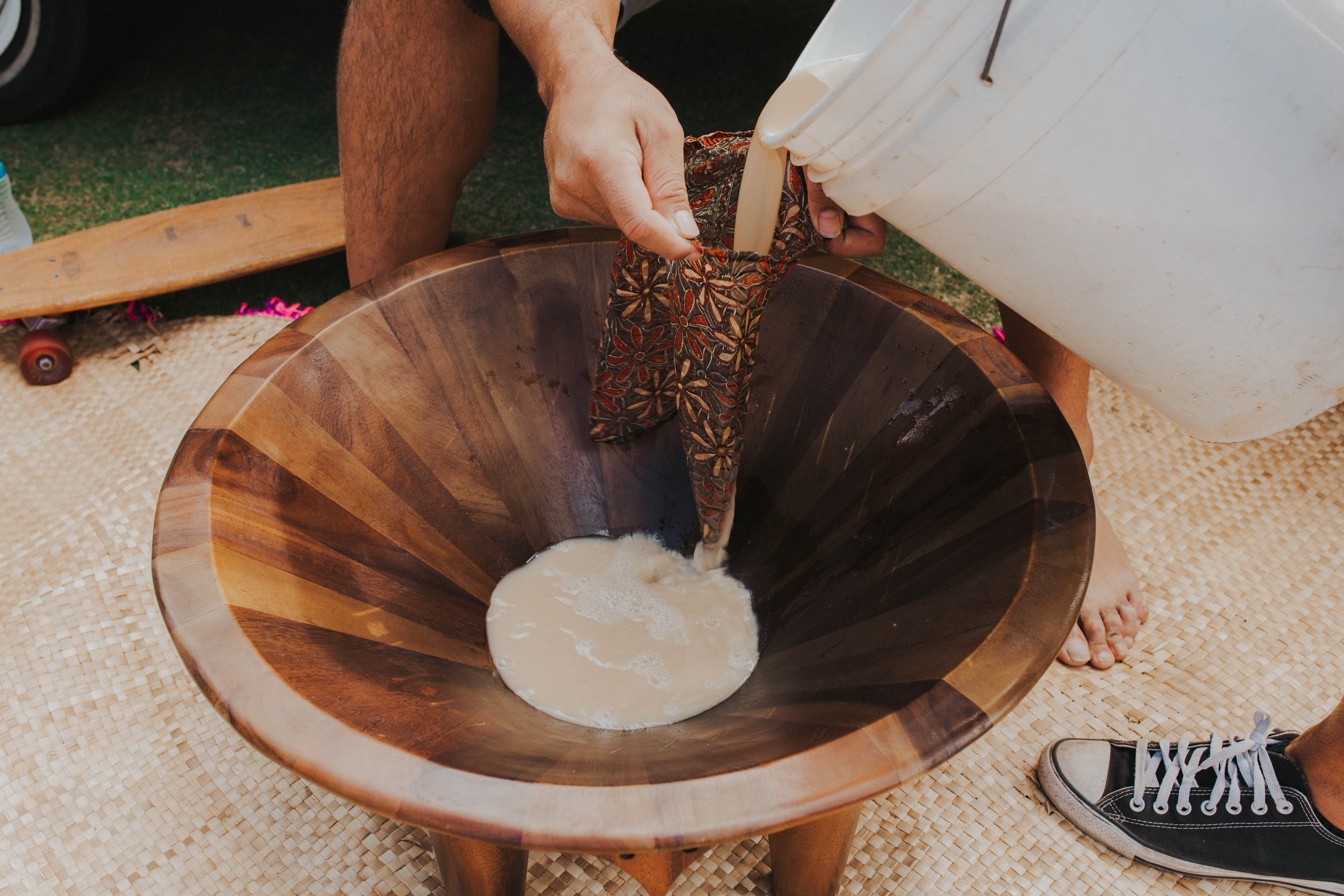 A person pours liquid from a white bucket through a cloth strainer into a large wooden bowl.