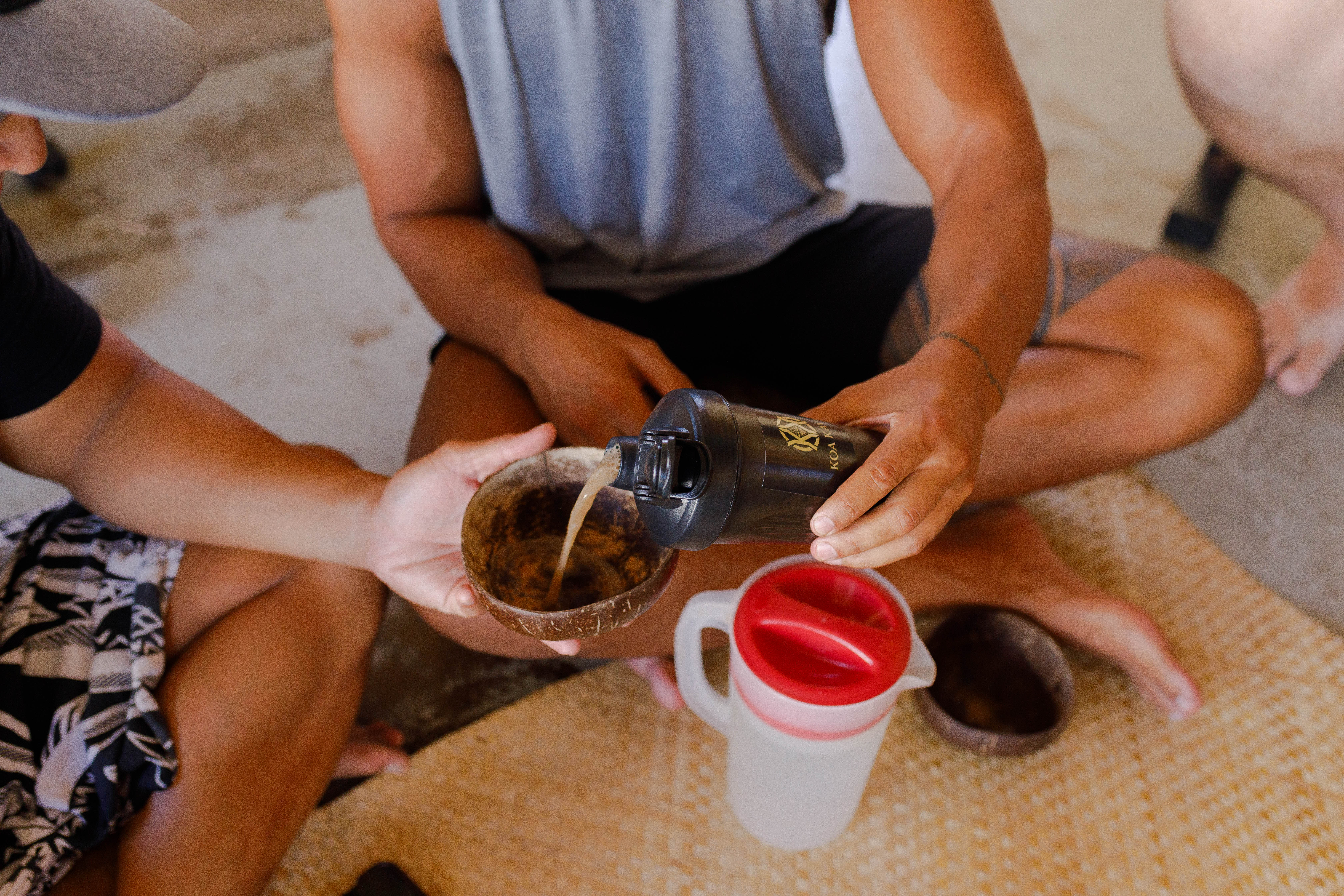 A person pours a drink from a black shaker bottle into a coconut shell bowl held by another.