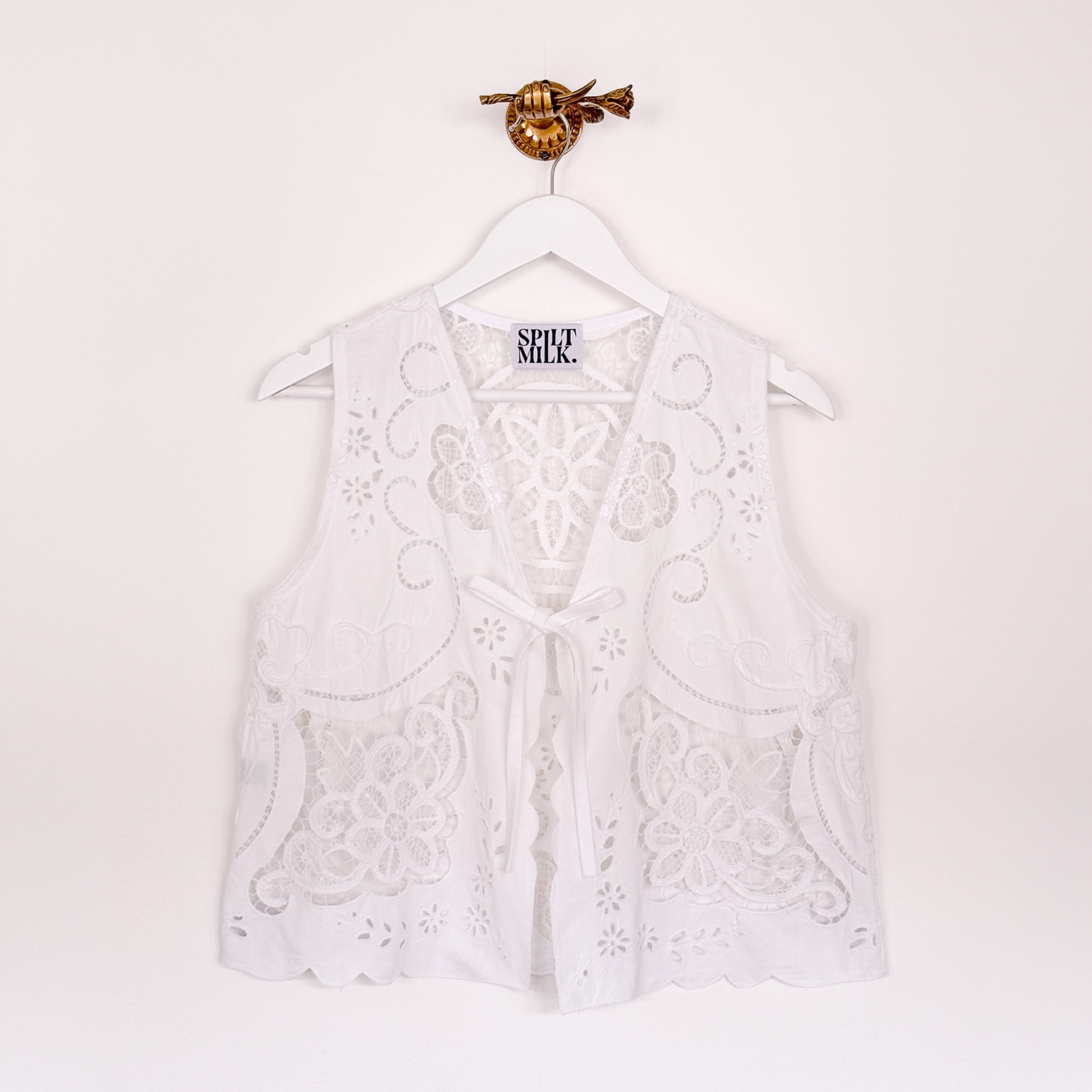 A white sleeveless lace vest with a floral pattern and tie-front hangs on a white hanger.
