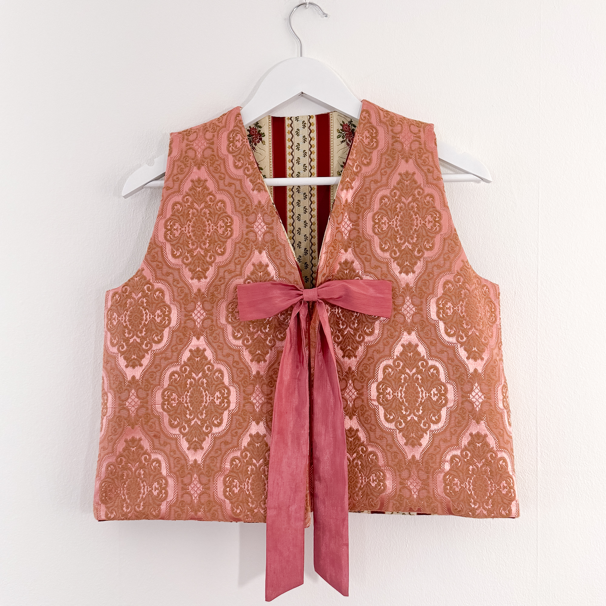 A pink and brown patterned vest with a pink ribbon tie hangs on a white hanger.