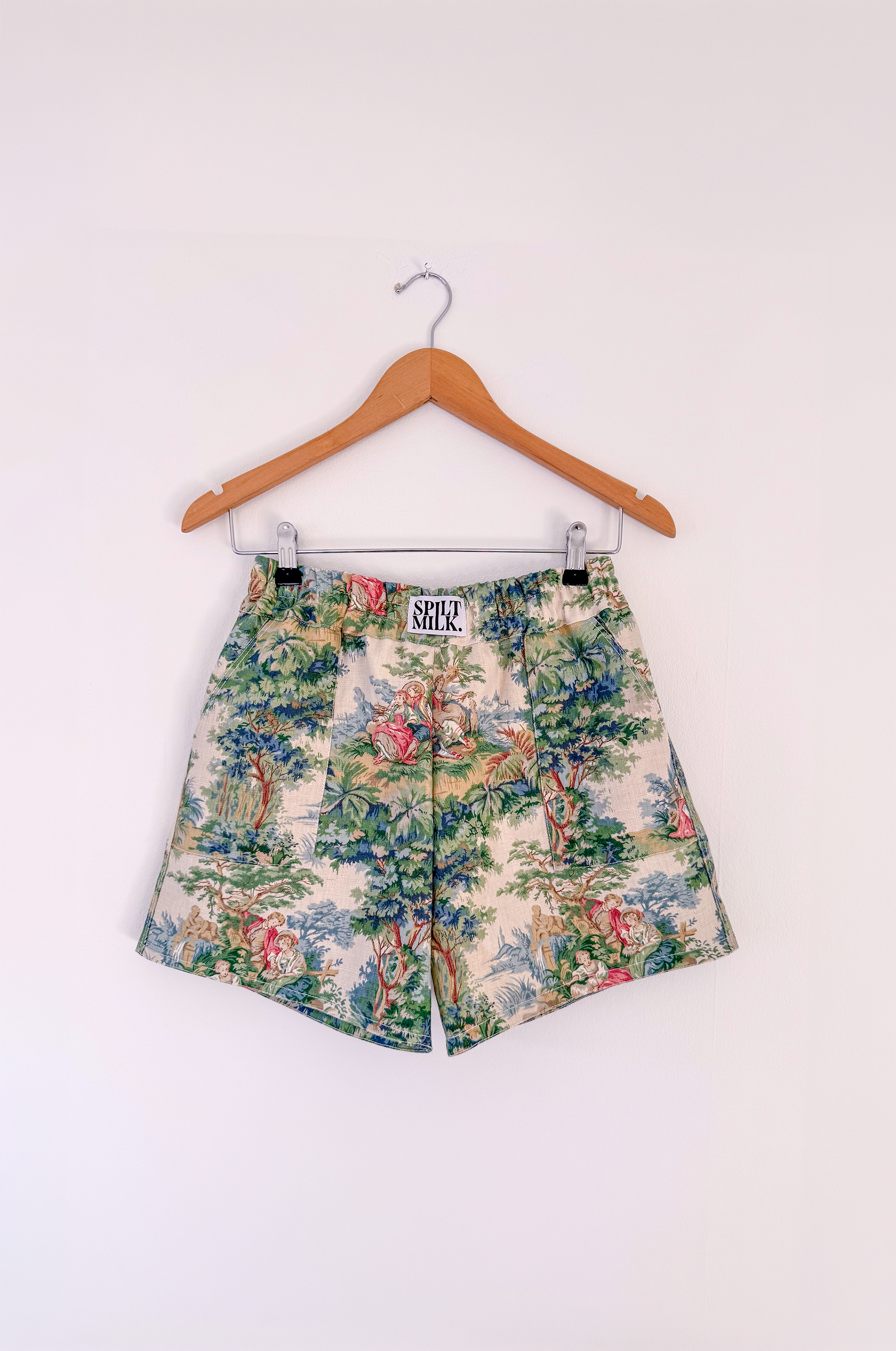A pair of shorts with a pastoral toile pattern hanging on a wooden hanger against a white wall.