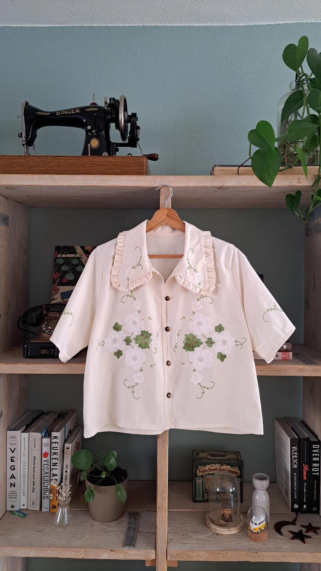 Vintage sewing machine and floral shirt on a wooden shelf with books and plants.