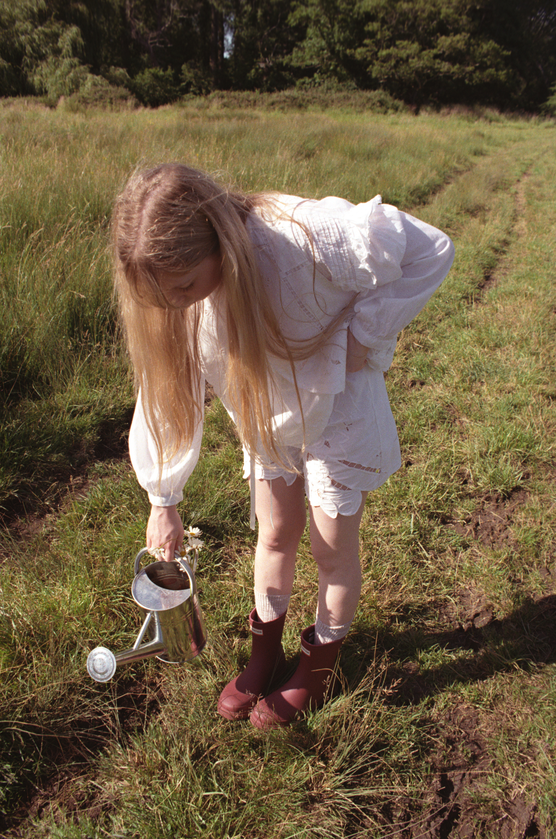 A person with long blonde hair in a white outfit holds a watering can in a grassy field.