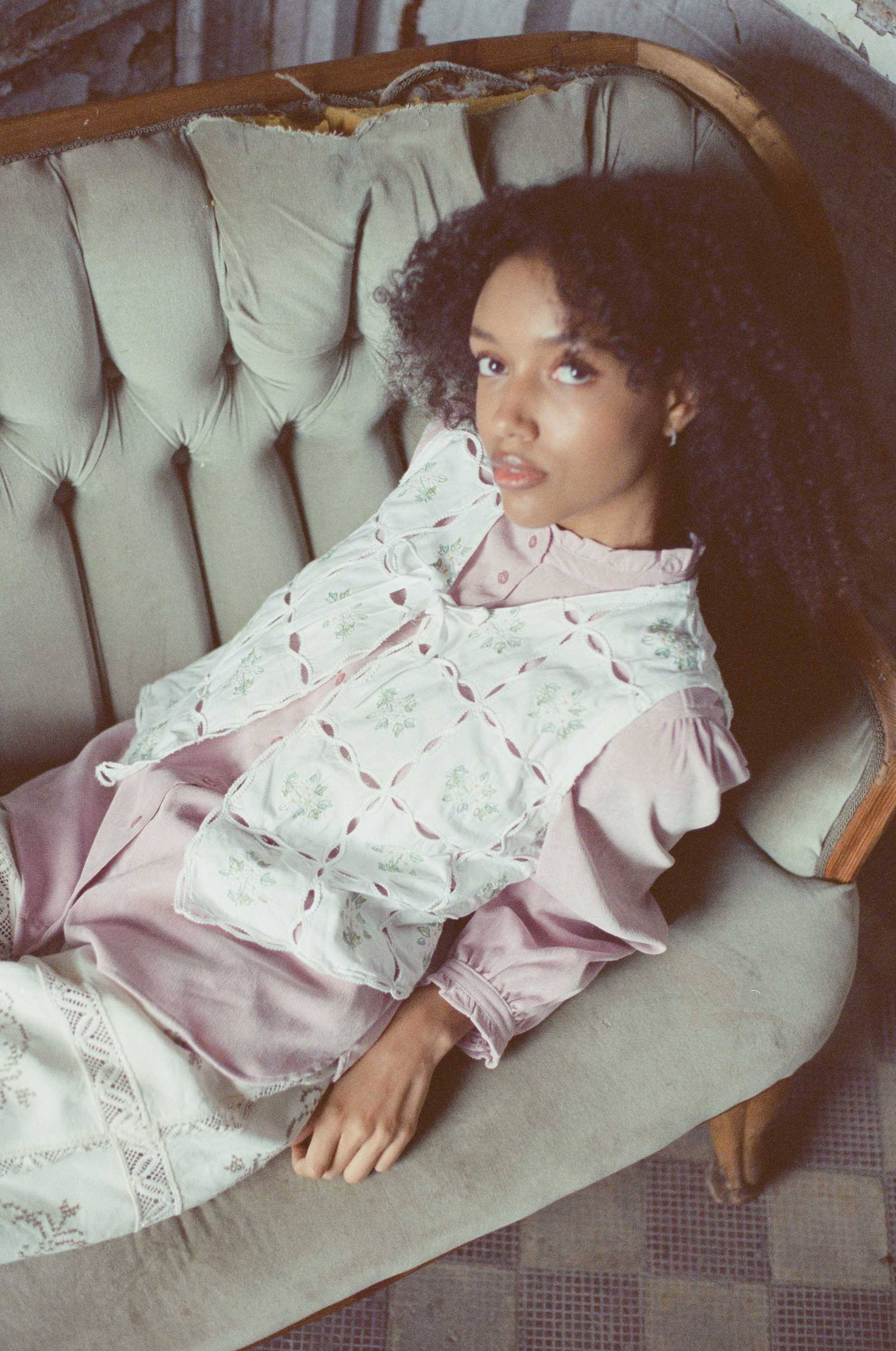 A person with dark curly hair reclines on a worn, tufted sofa, looking toward the camera.