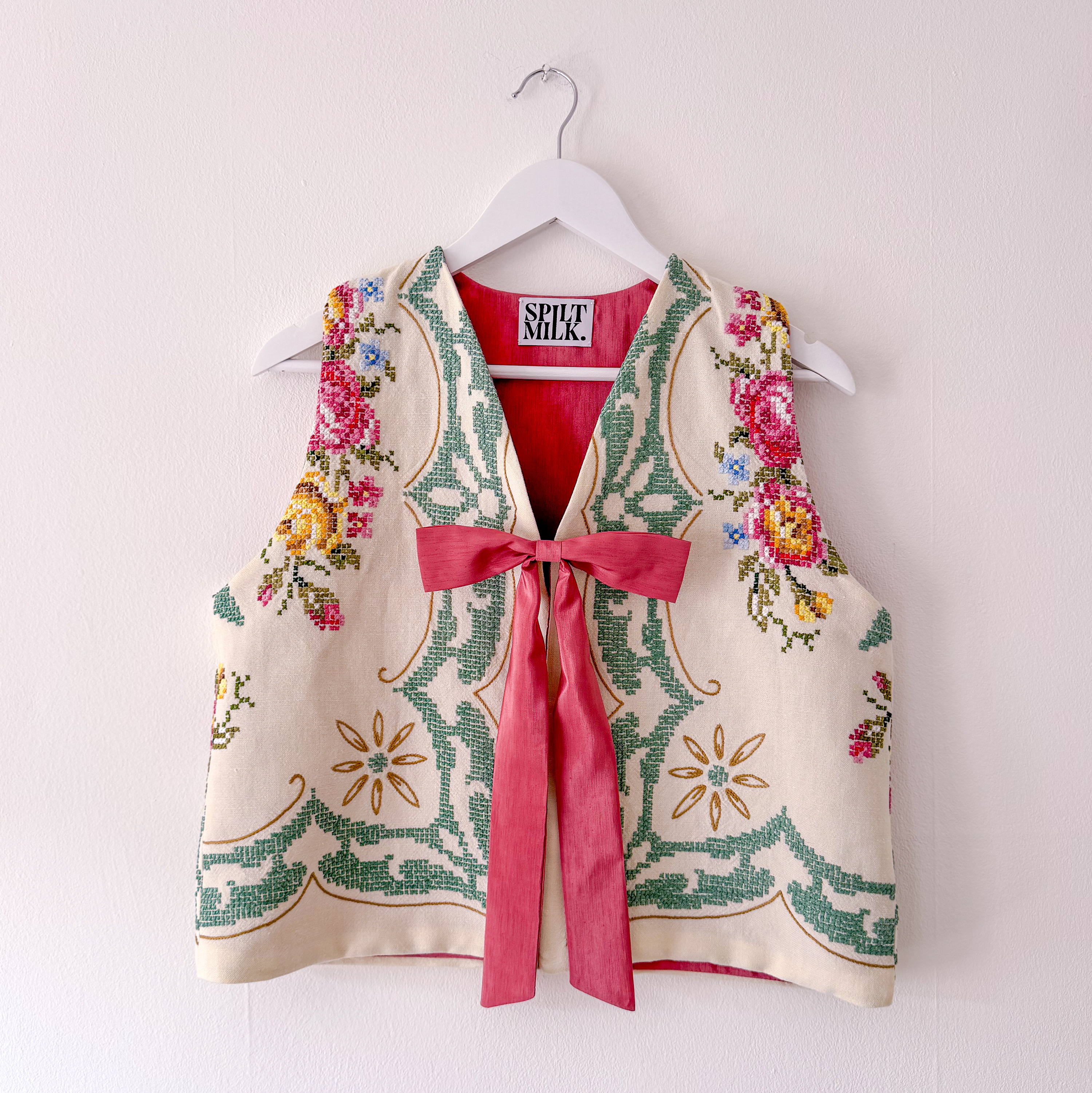 A cream-colored, cross-stitched vest with floral embroidery and a pink ribbon tie, hanging on a white hanger.
