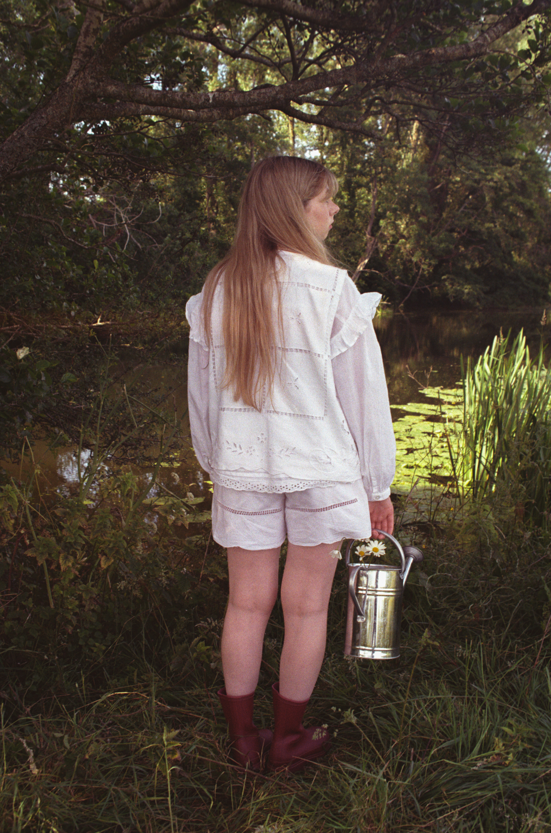 A person in a white outfit and red boots stands in a field holding a watering can.