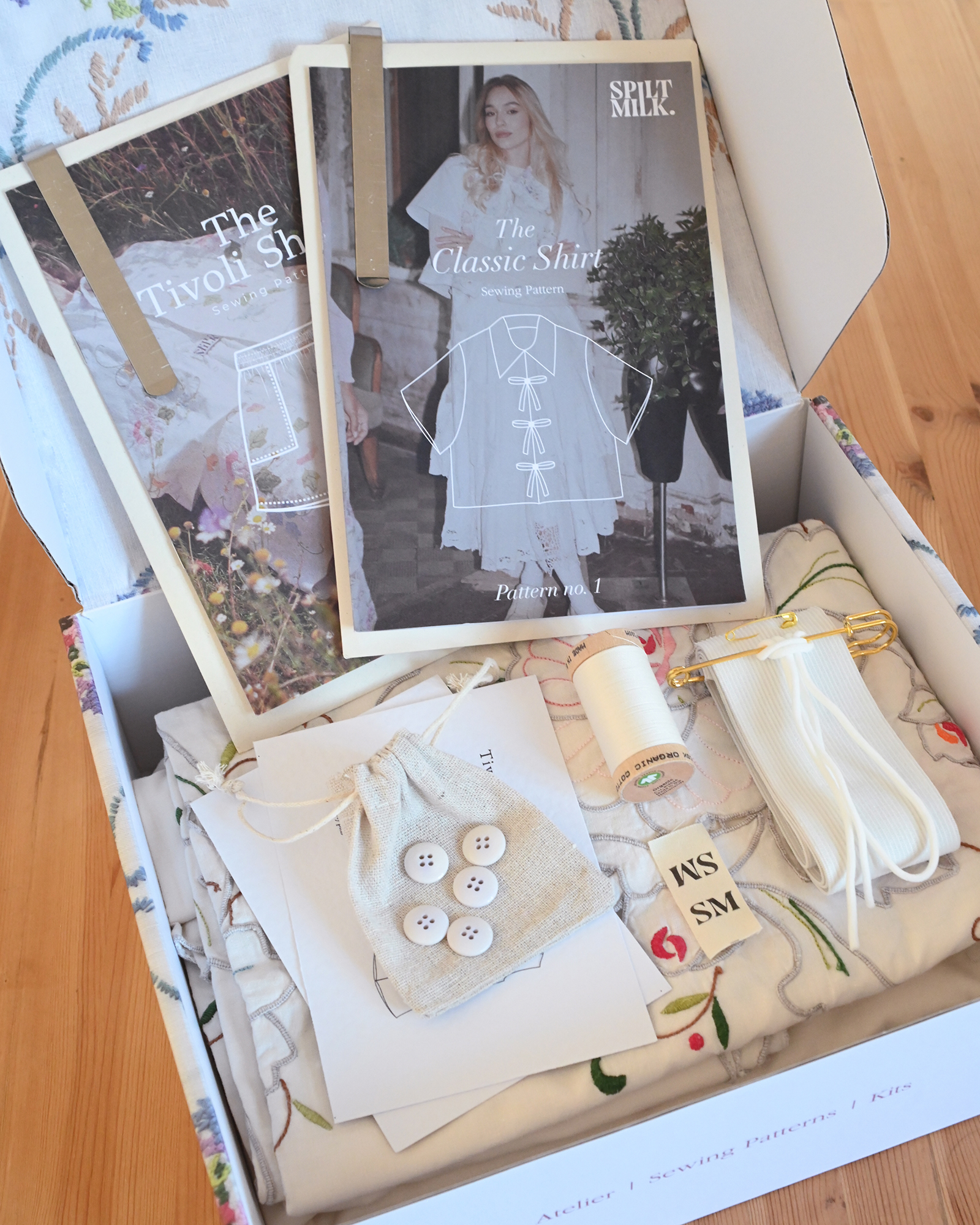 An open box containing a sewing kit with patterns, embroidered fabric, buttons, and a spool of thread.