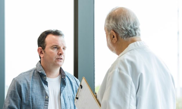 Two men talking, one holding a clipboard, near a window.