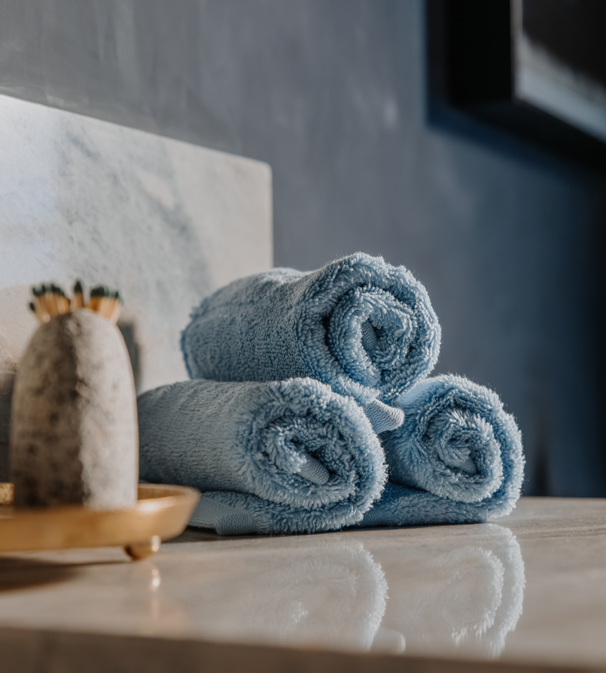 Three light blue towels rolled up and stacked on a reflective countertop next to a decorative object.