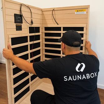 A technician in a Saunabox t-shirt assembles the wooden panels of an indoor sauna.
