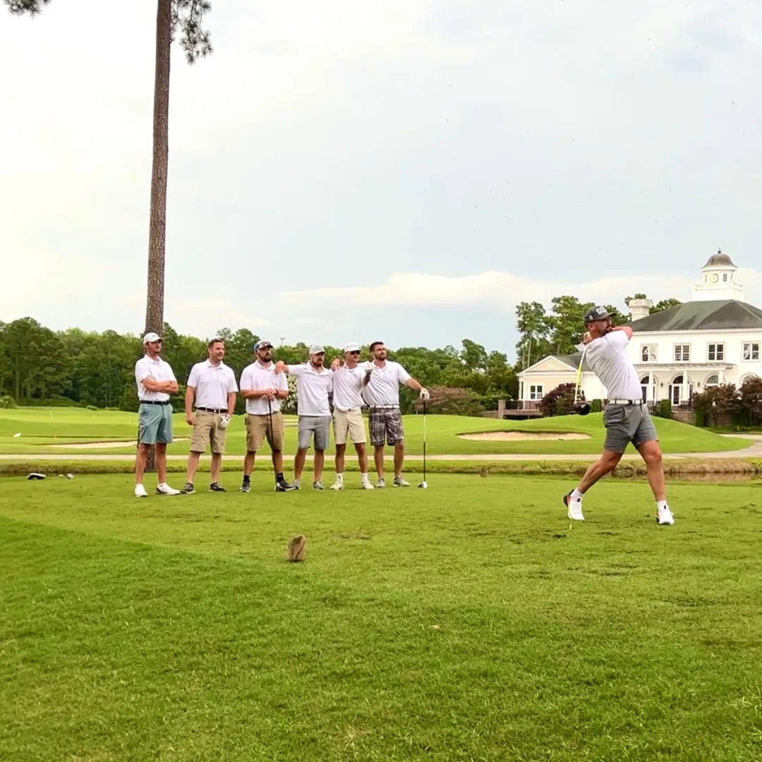 Group of men in white shirts watching a golfer swing on a golf course.