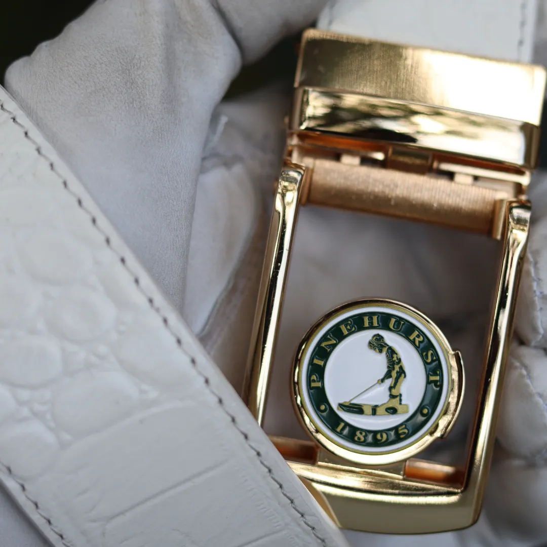 Gold belt buckle with 'Pinehurst 1895' logo, held by a gloved hand.