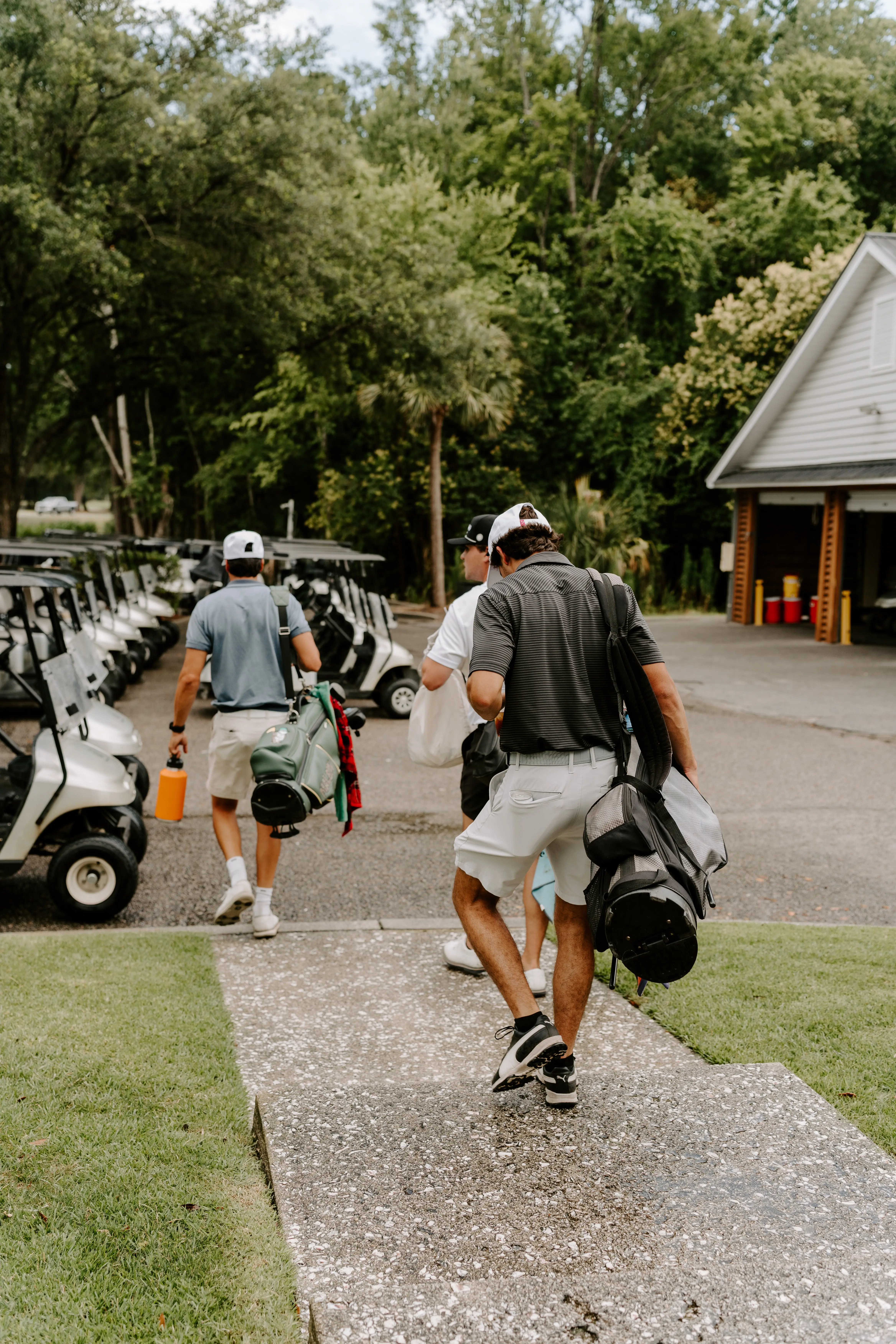 Three people walking with golf bags near parked golf carts and trees.