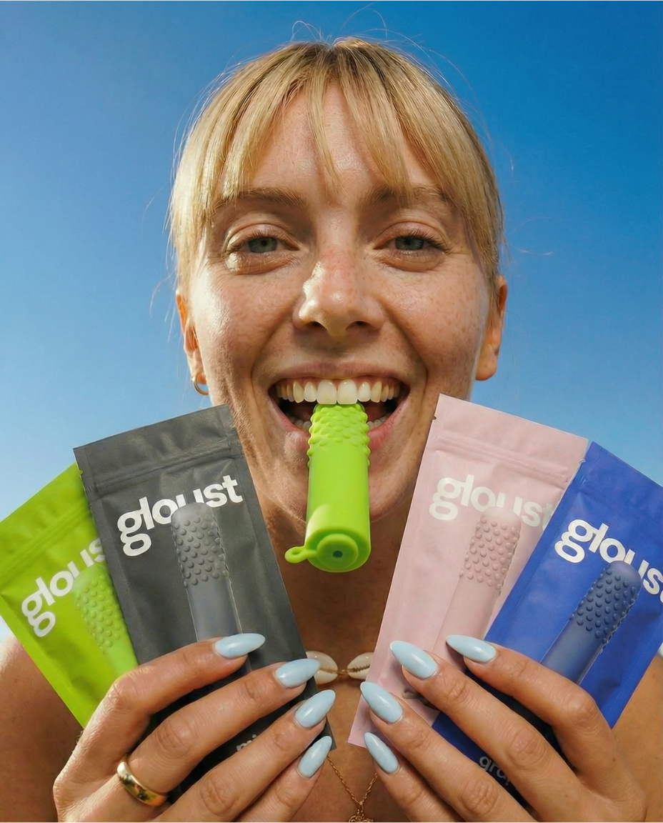 A smiling woman holds colorful 'gloust' brand pouches and bites a green textured object against a blue sky.