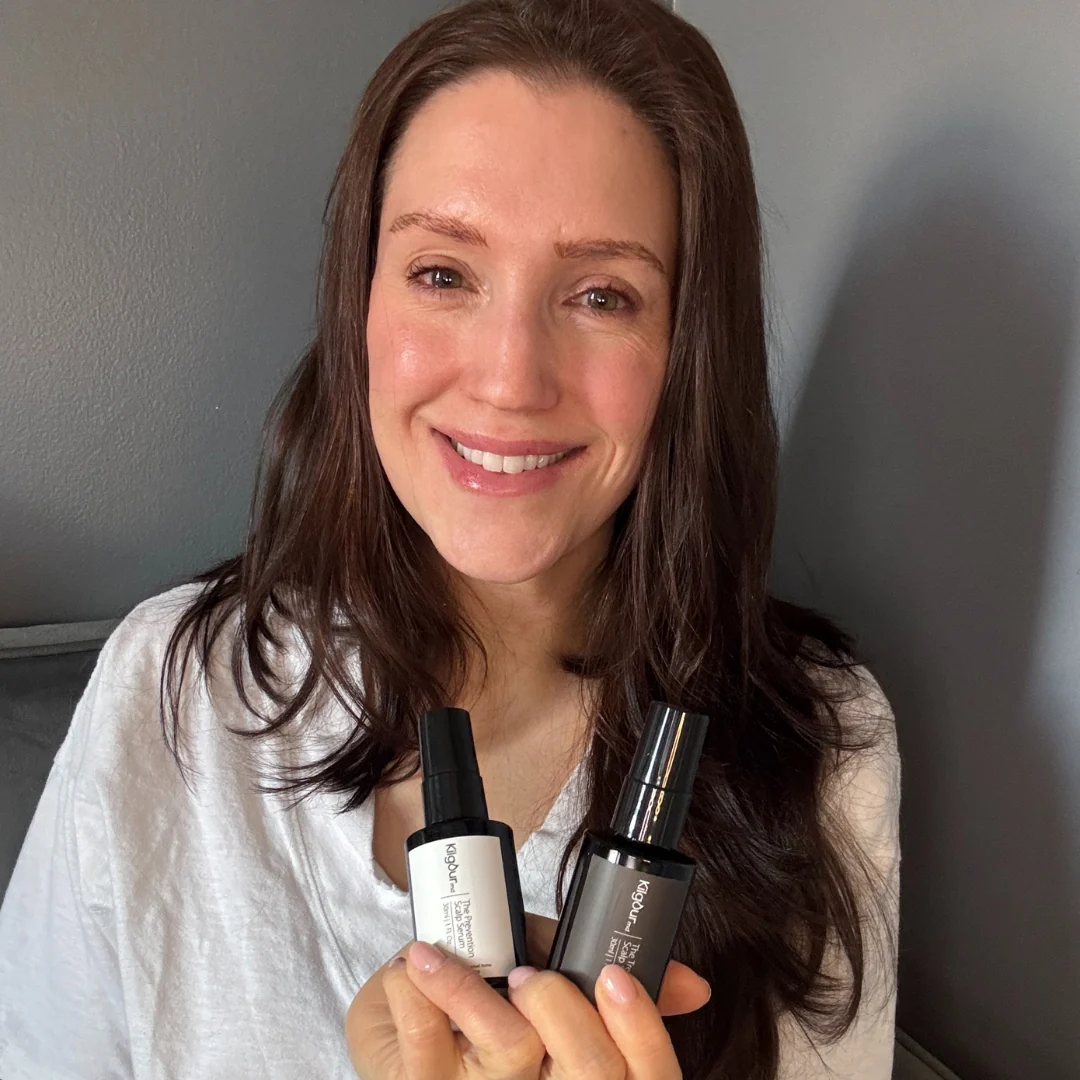 Person smiling, holding two skincare bottles against a gray background.